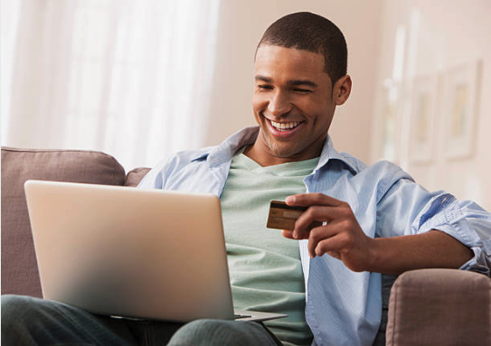 A man sits on the couch with his laptop and a credit card in his hand