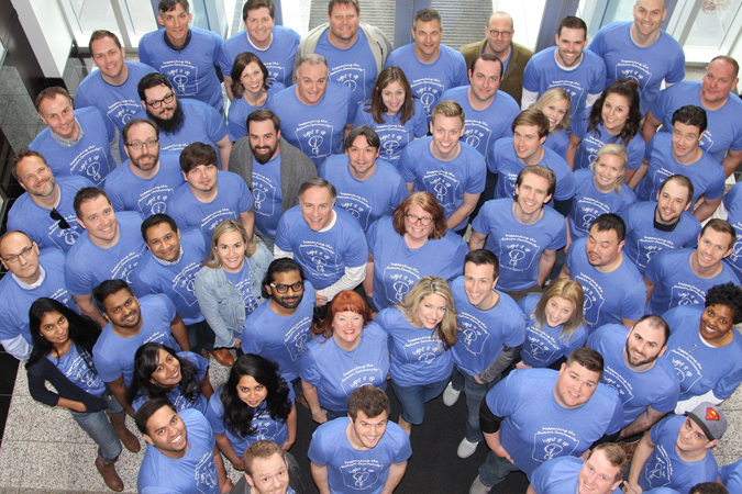 A group of Rx Savings Solutions employees in matching blue shirts looks up at a camera positioned above them in the company lobby