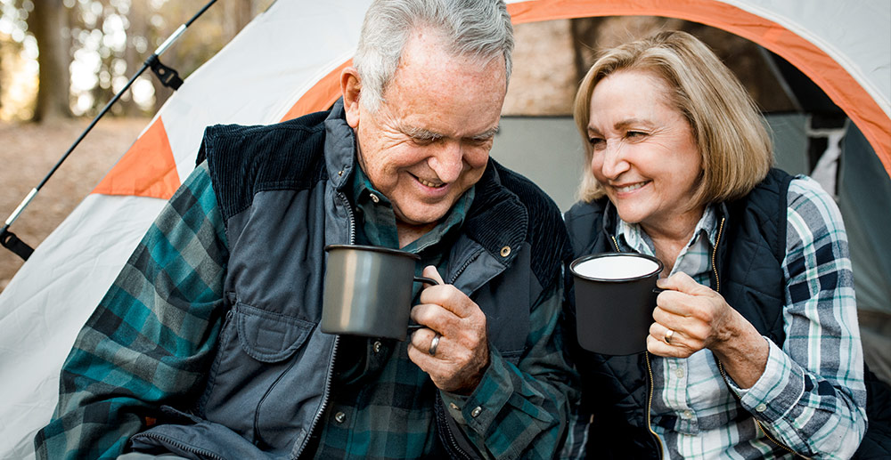 An older man and woman sit outside of a camping tent drinking out of coffee mugs