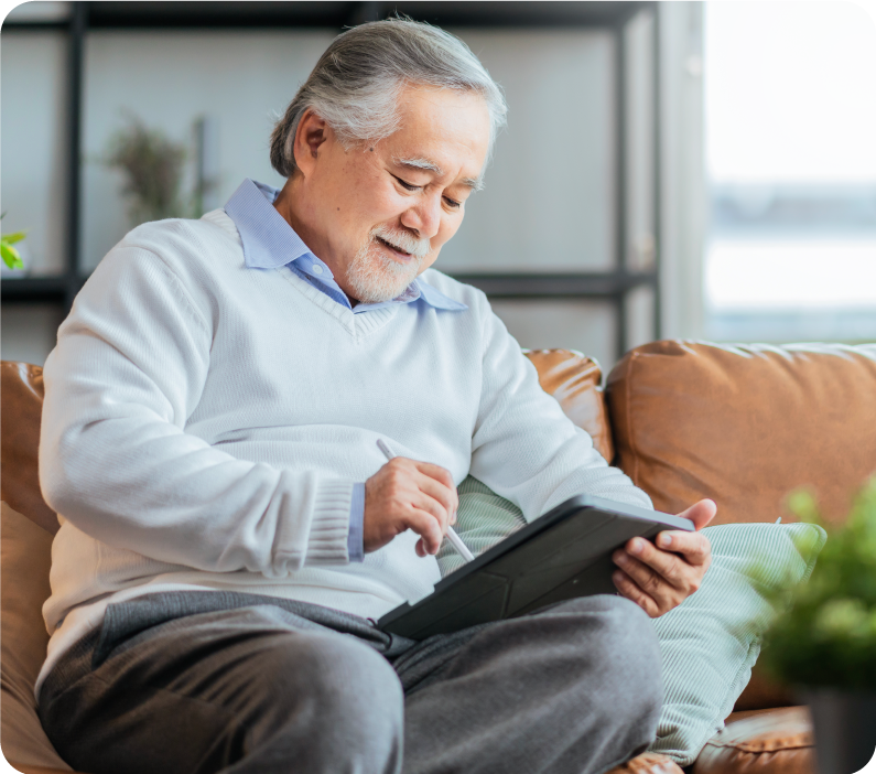 An older man sits on his couch reading from a tablet device.